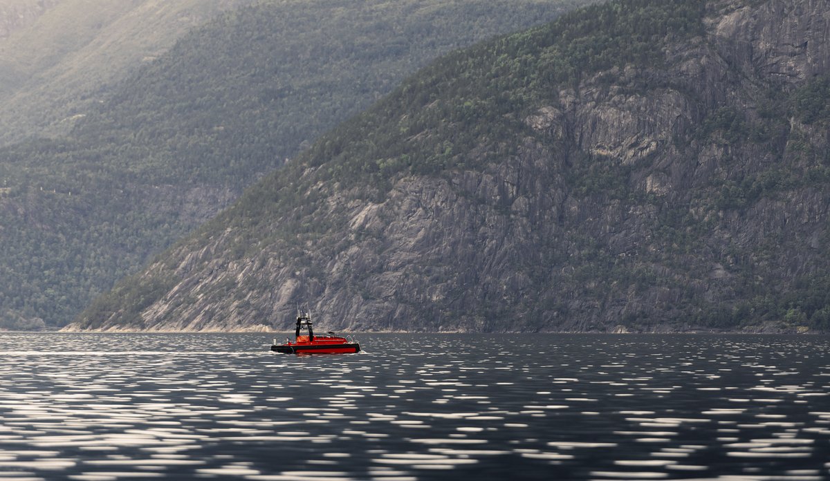 
Oransje fartøy ute på fjord med bratte fjellsider rundt.