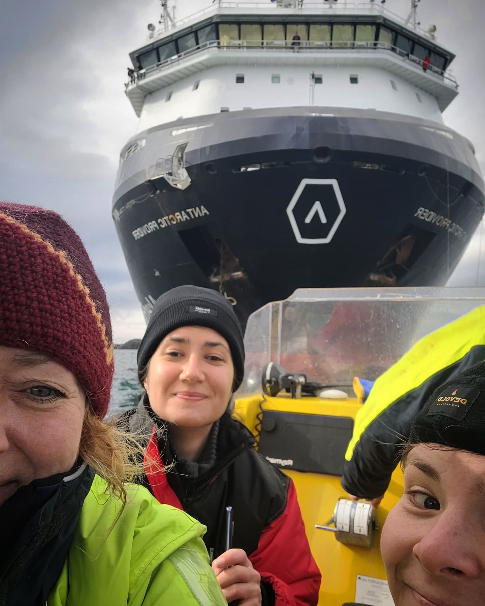tre kvinner i lettbåt på sjøen med stort cargoskip bak