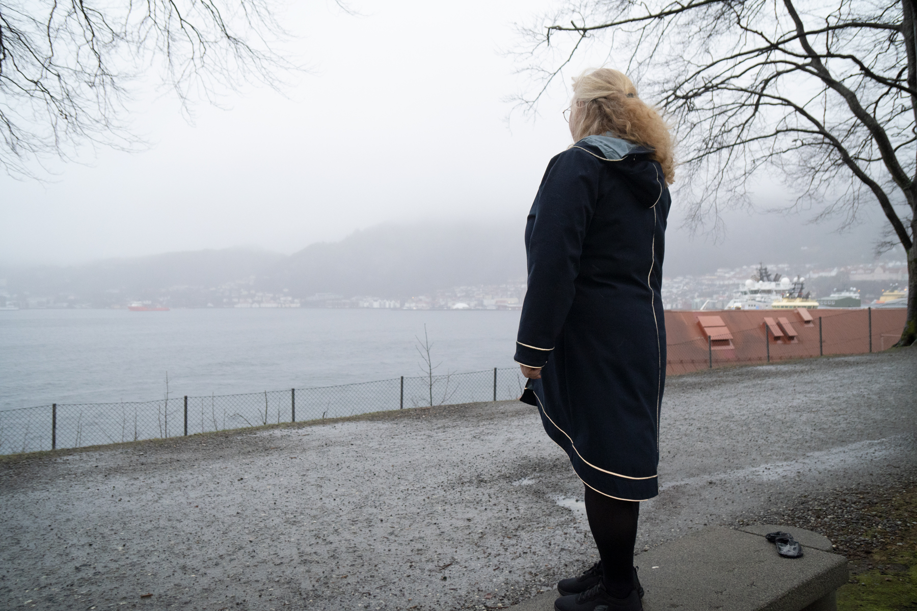Kvinne i mørk frakk står med ryggen til og ser ut mot sjøen, grått regnvêr.