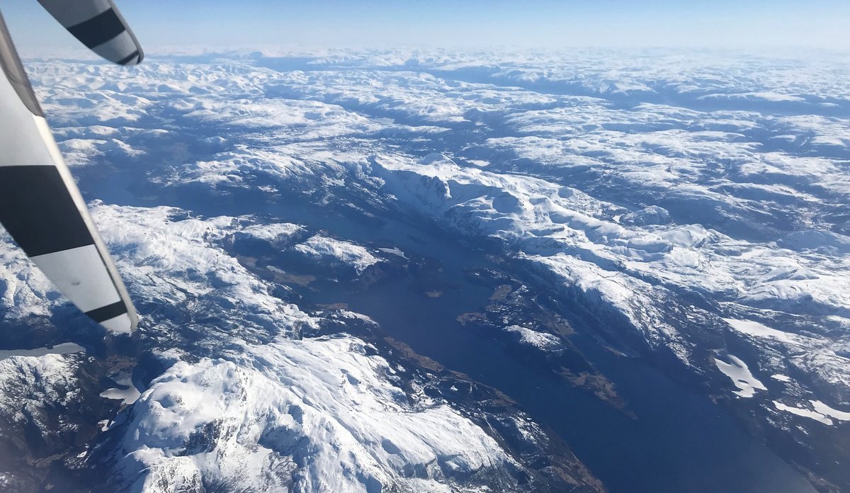 Fjord omgitt av snøkledde fjell sett ovenfra fra et fly med deler av propell i forgrunnen.