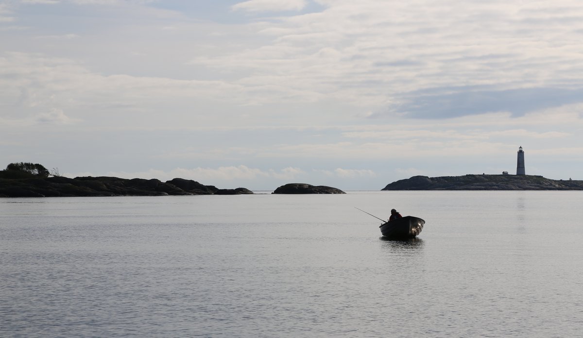 
En person fisker fra en robåt som ligger på sjøen. I horisonten ser en noen holmer, og på den siste av dem står et fyr. 