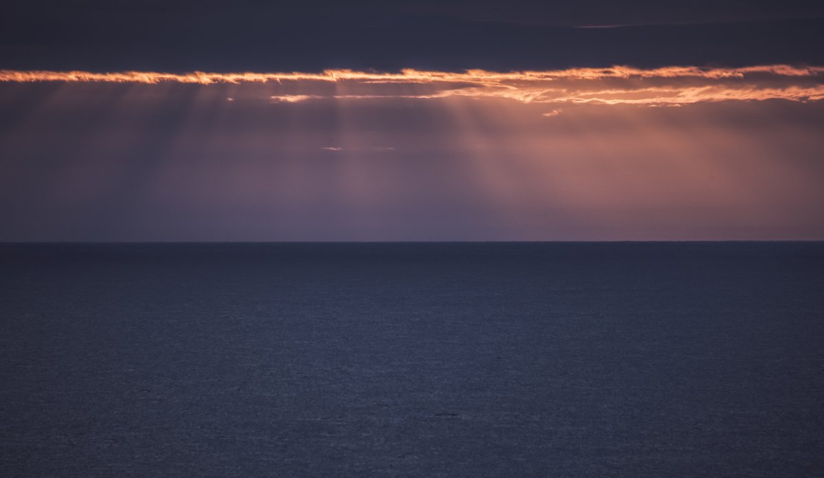 
Blikkstille hav, horisont i bakgrunnen og solstråler som kommer gjennom skylaget