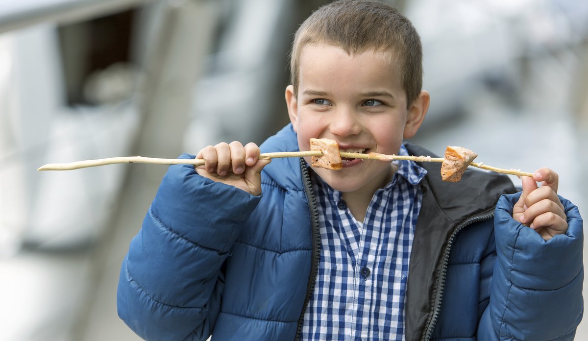 
Liten gutt med blå jakke spiser grillede lakseterninger tredd på en pinne.