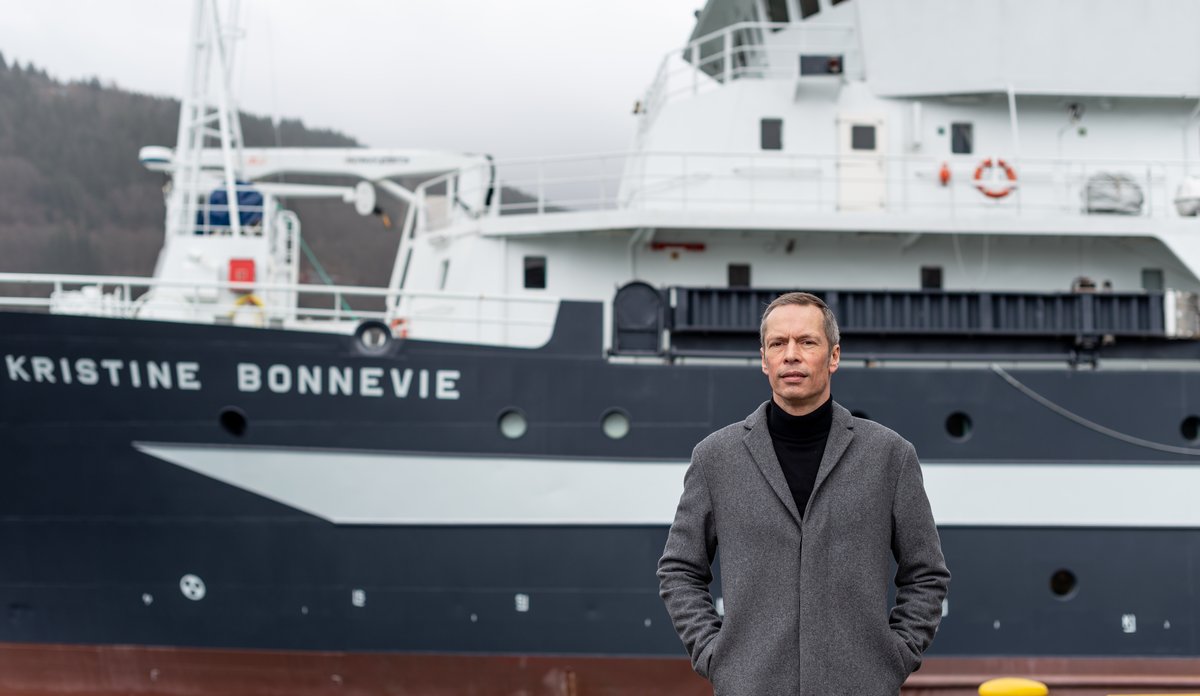 Mann i grå kåpe foran forskningsskip