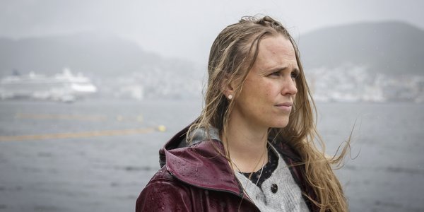 

Kvinne med lilla regnjakke og vind og regn i håret, med en fjord, et cruiseskip og bebyggelse i bakgrunnen.