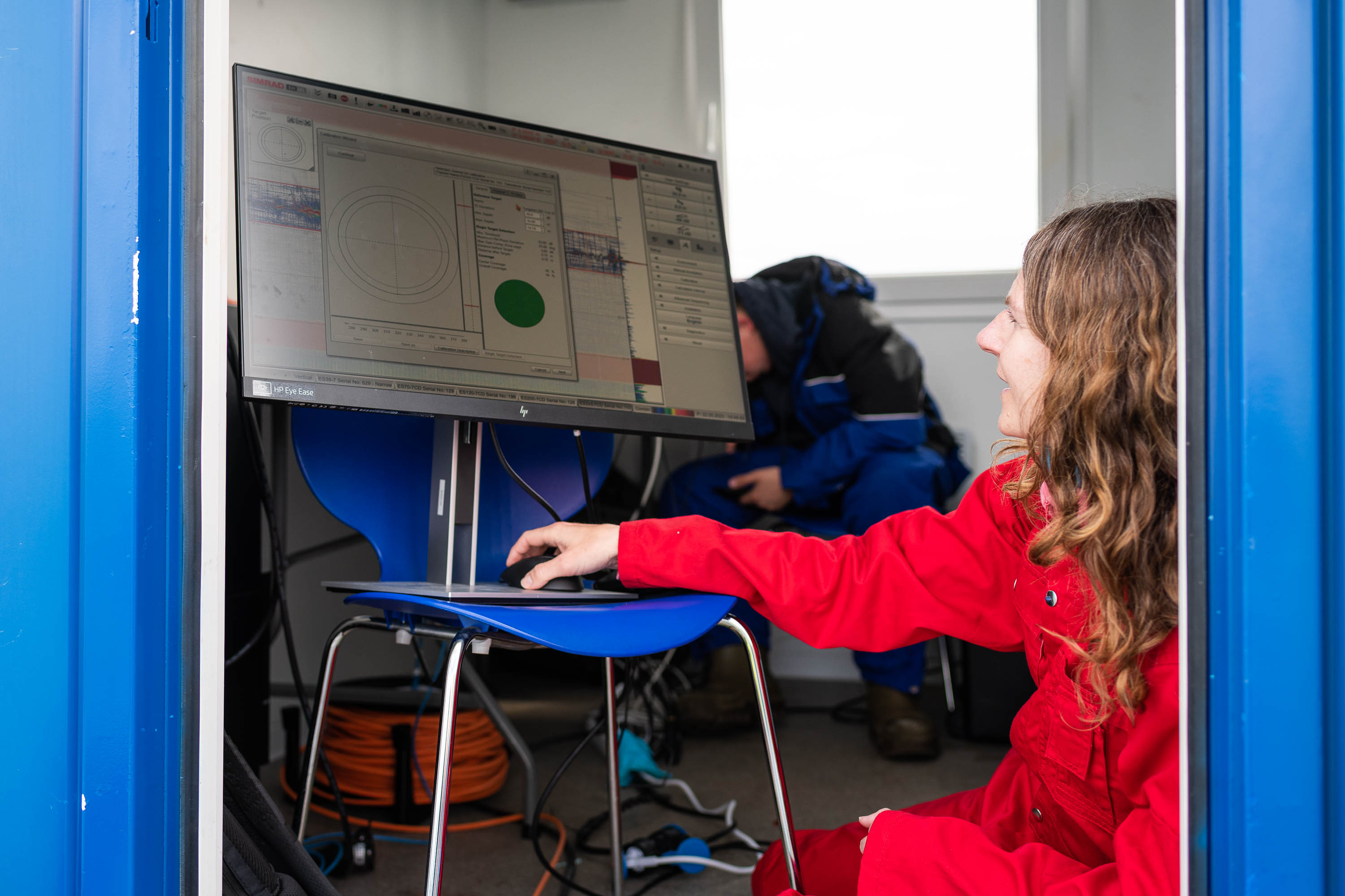 Ei kvinne i raud kjeledress sit på hug framfor ein dataskjerm. Dataskjermen står på ein stol, rett innanfor døra i ein kontainer.