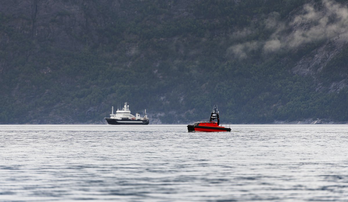 Foto av en fjord med forskingsfartøy og liten båt-liknende drone i forgrunnen, i en fjord.
