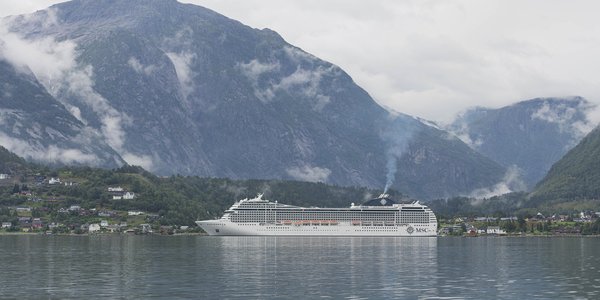 

Hvit cruiseskip i en fjord med skydekkede fjell i bakgrunnen.