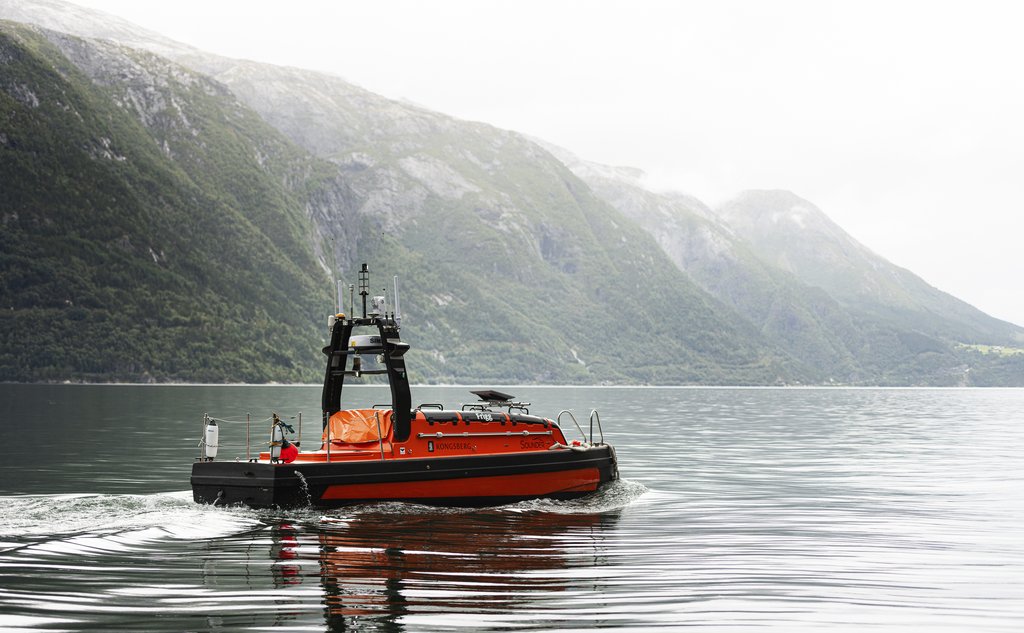 Oransje fartøy ute på fjord med bratte fjellsider i bakgrunnen