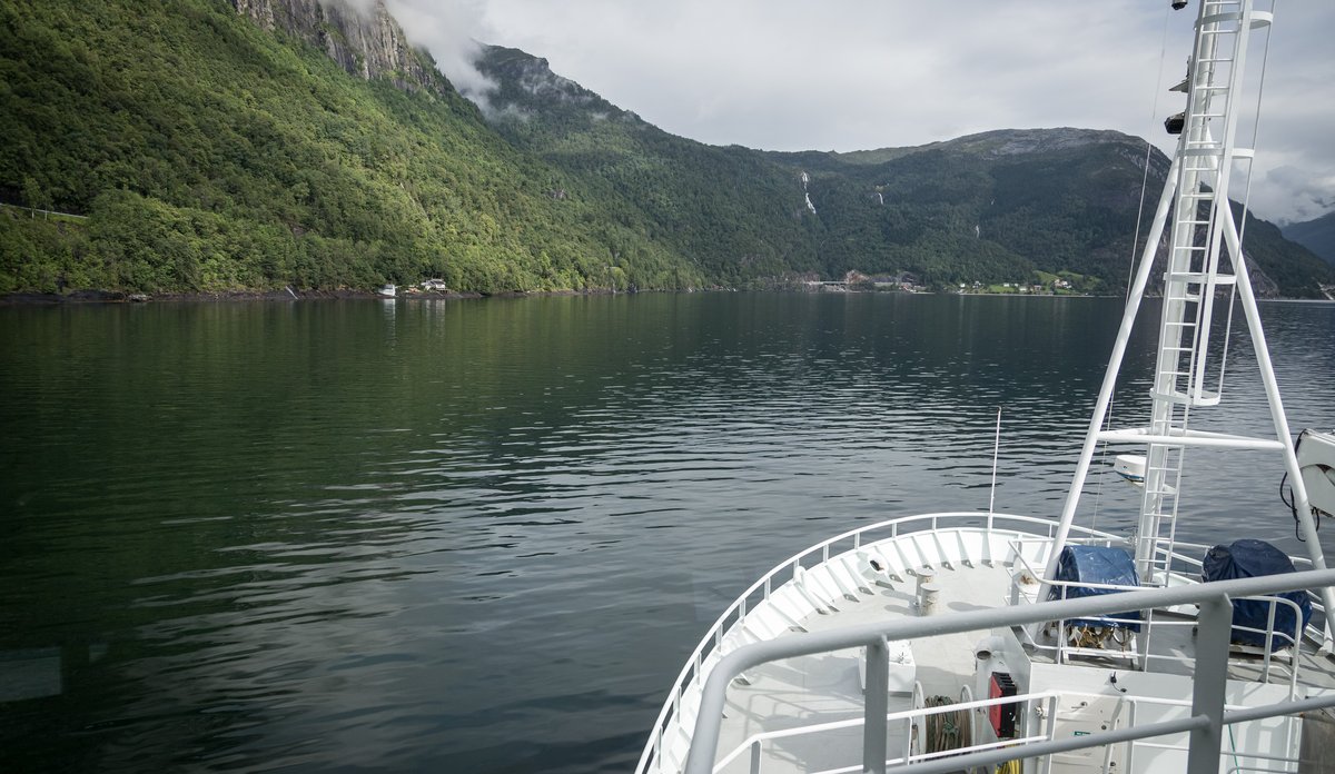 
Bilde av baugen på en båt som er på vei innover en fjord. Sjø foran båten, fjell på siden.