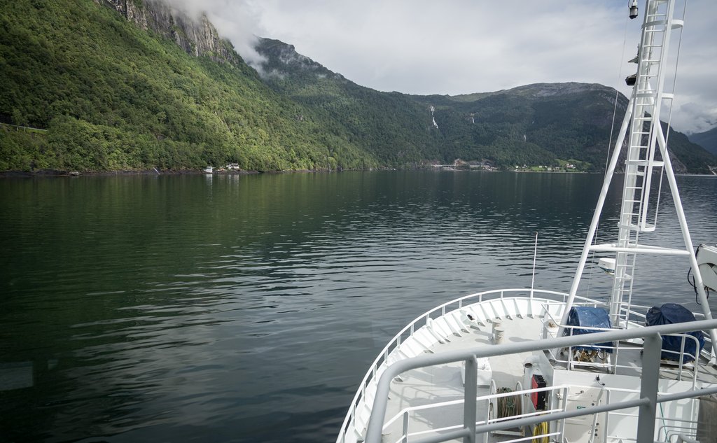 Bilde av baugen på en båt som er på vei innover en fjord. Sjø foran båten, fjell på siden.