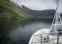 Bilde av baugen på en båt som er på vei innover en fjord. Sjø foran båten, fjell på siden.