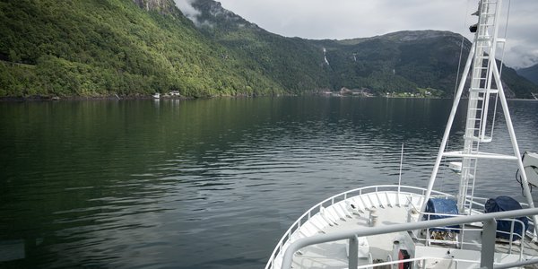 

Bilde av baugen på en båt som er på vei innover en fjord. Sjø foran båten, fjell på siden.