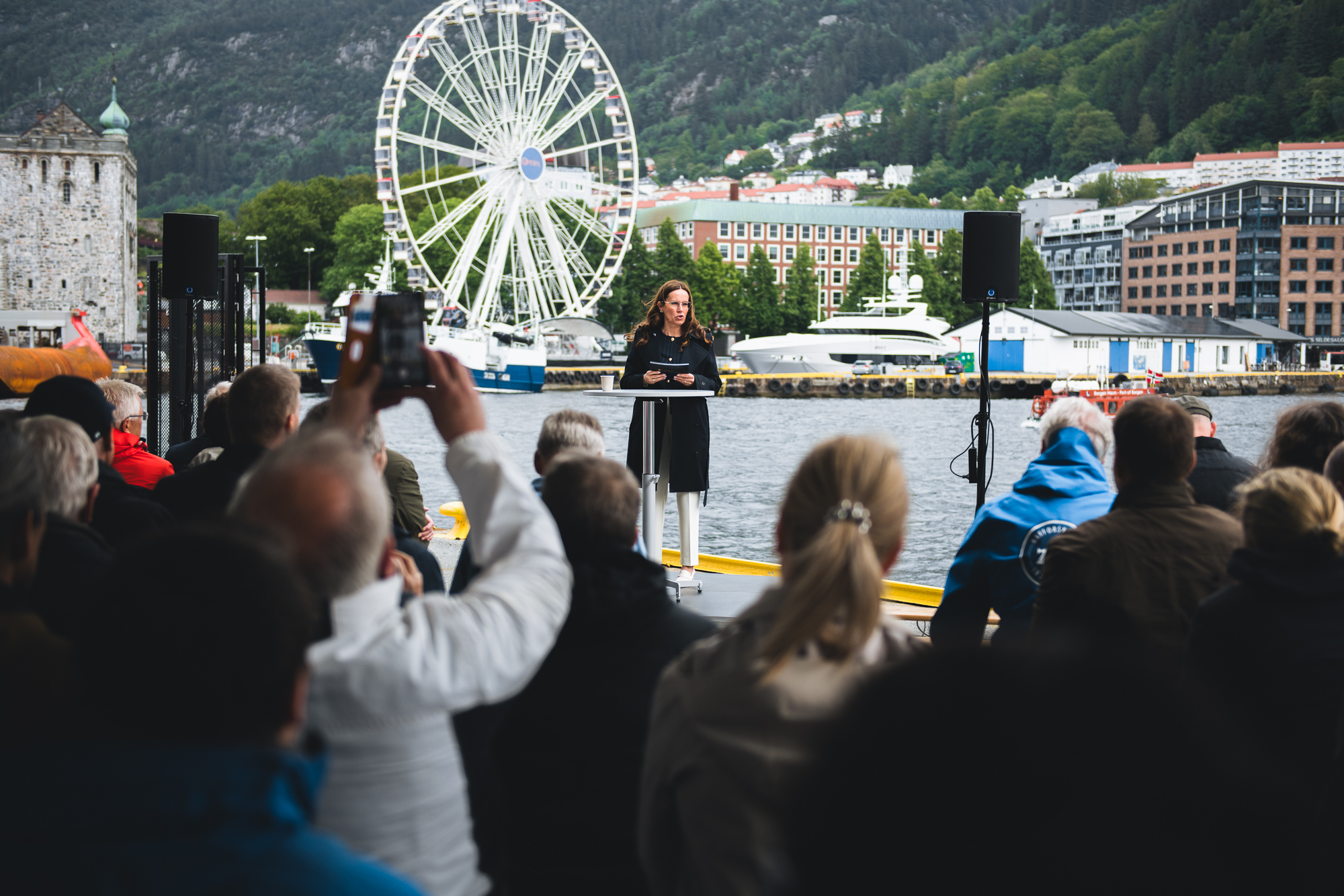 Kvinne på podium med mennesker i forgrunnen og bybilde med Vågen og Bryggen i bakgrunnen.