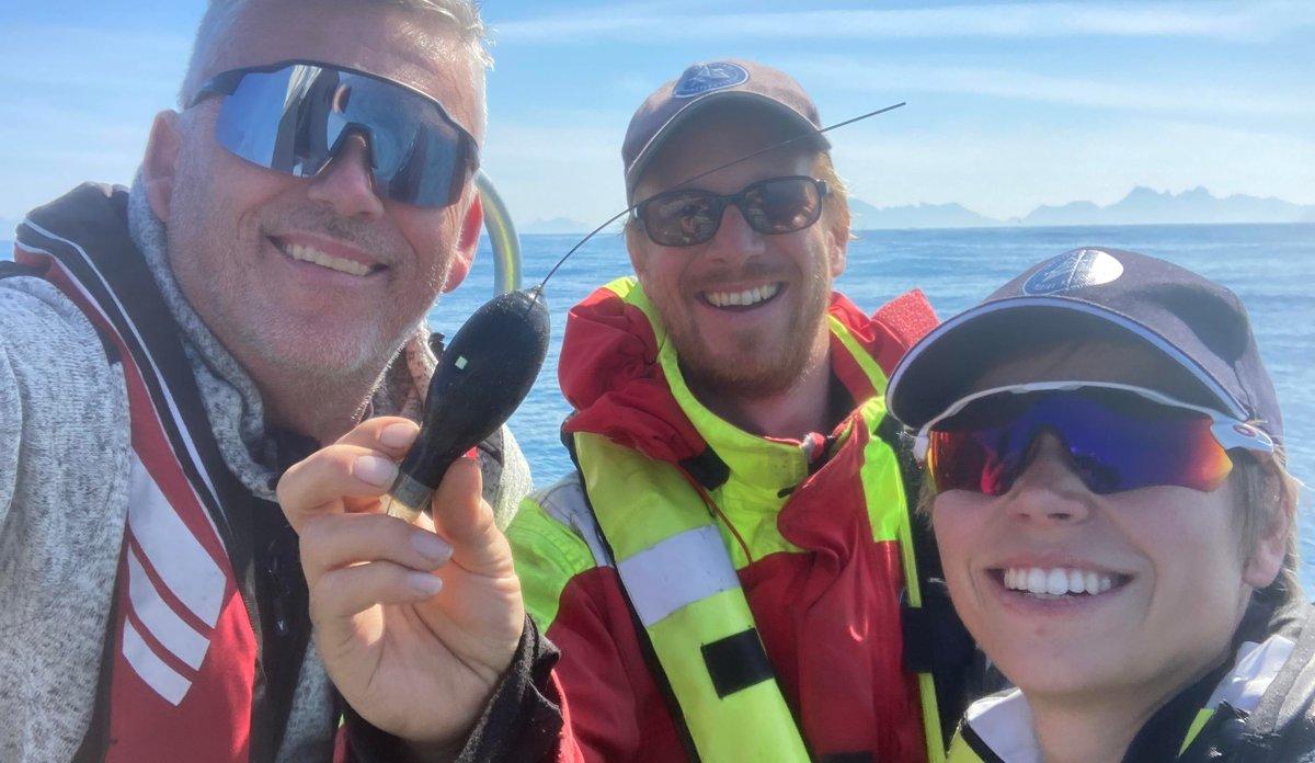 Tre personar smiler mot kameraet. Dei har alle på seg solbriller og redningsvest. Personen i midten held opp eit lite satellittmerke, som han viser til kamerat.