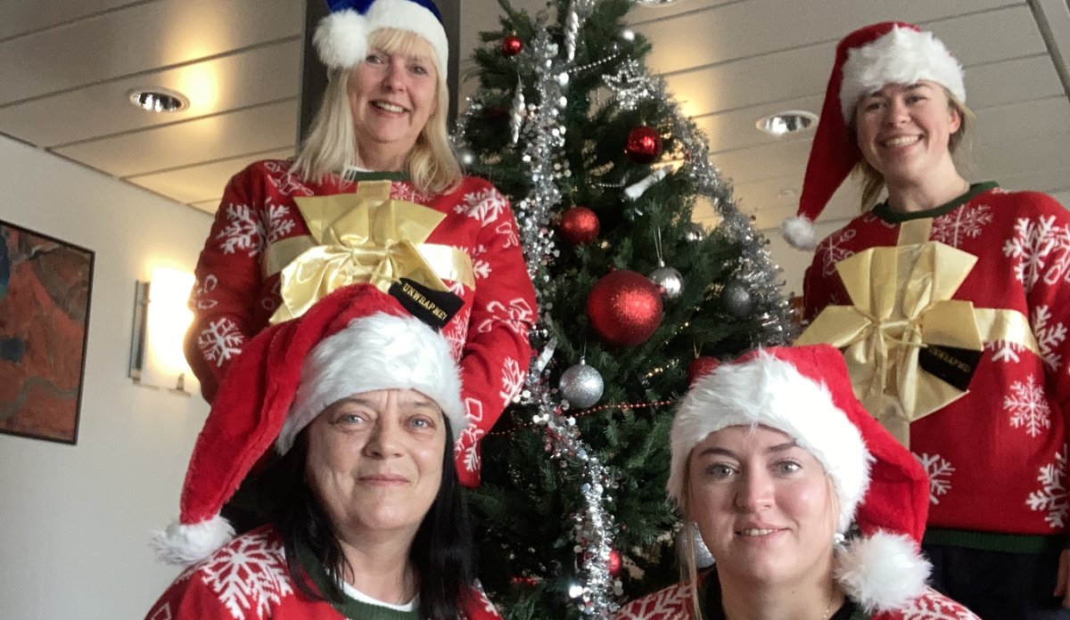 Fire kvinner med røde julegensere og nisseluer og et juletre i bakgrunnen.