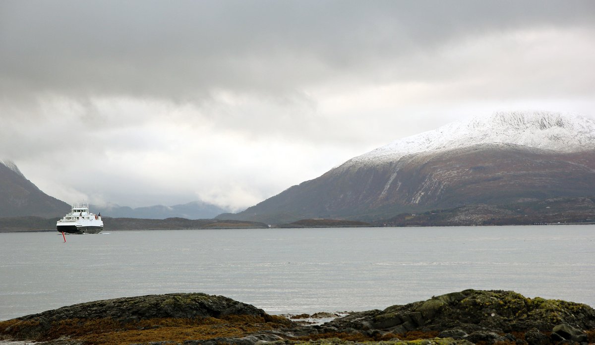 
Fjord med ferje og snøkledde fjell og grå skyer i bakgrunnen