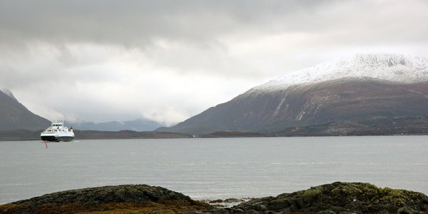 

Fjord med ferje og snøkledde fjell og grå skyer i bakgrunnen