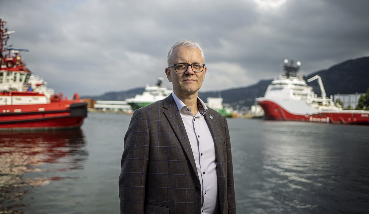 
En mann med briller og i dressjakke står mot kameraet. i bakgrunnen skimter en havet og noen båter