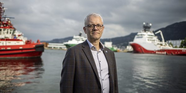 

En mann med briller og i dressjakke står mot kameraet. i bakgrunnen skimter en havet og noen båter