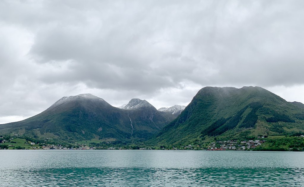 Bygd som ligger nedenfor fjeller med en fjord i forgrunnen under grå skyer