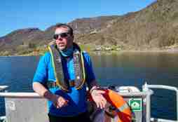 Prosjektleiar Rune Nilsen står på dekk på båten Emmy Egidius. Han har på seg redningsvest og solbriller. I bakgrunnen ser ein Etnefjorden, og den klare blå himmelen. 