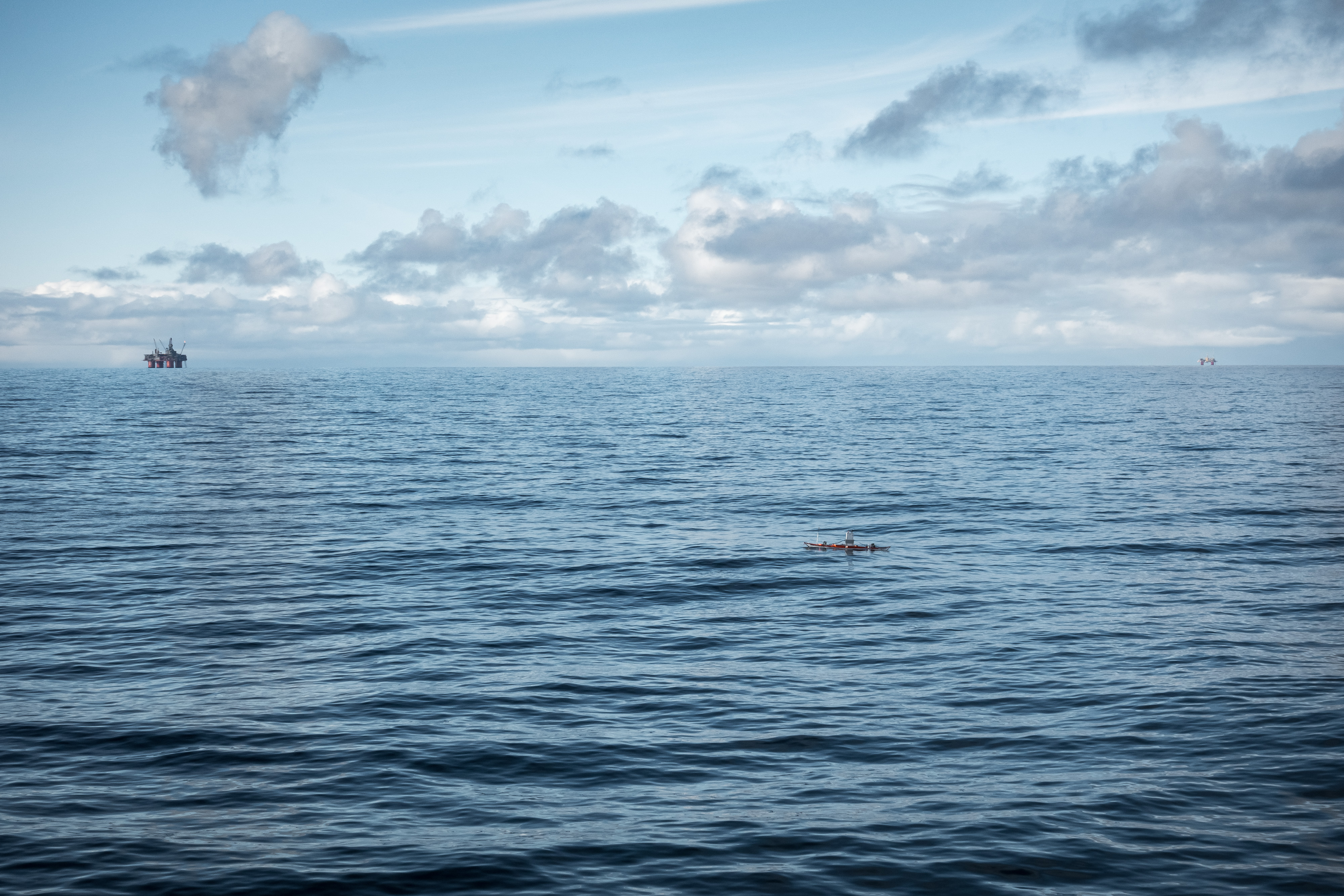 en liten kajakk i blått hav og strålende sol med to oljeplattformer i horisonten bak