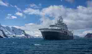 Forskningsfartøy på sjøen ved Antarktis, snødekte fjell i bakgrunnen.