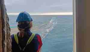 En kvinne i oljehyre og med hjelm på seg ser ut et vindu på en båt. Hun ser på ein trål som ligger i overflaten av havet.